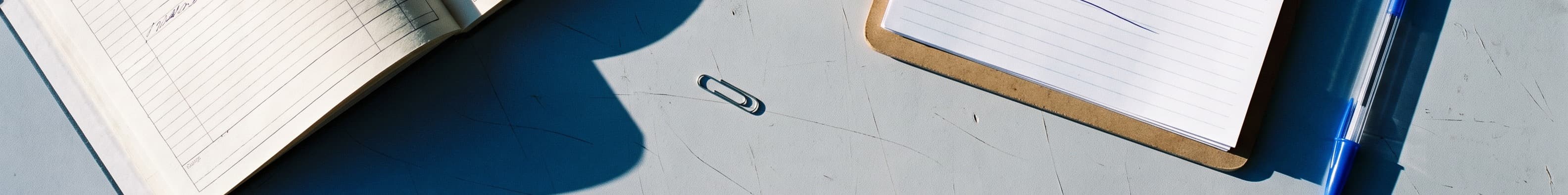 A ruled ledger open beside a clipboard, a paper clip and a pen on a desk.